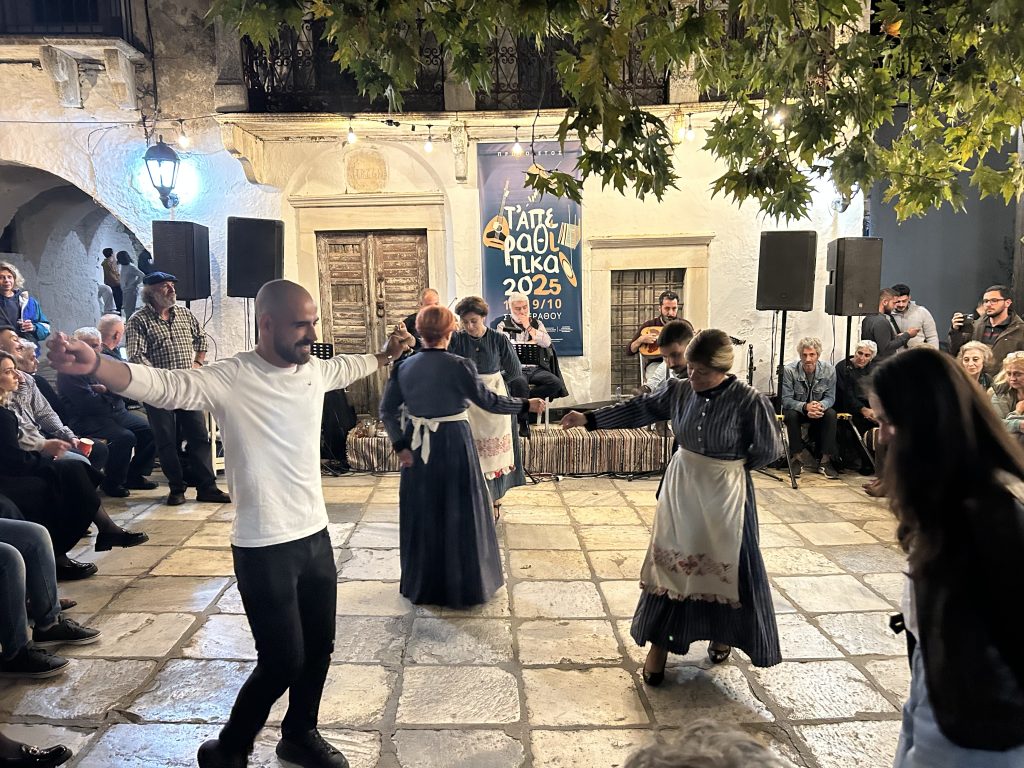 Local residents in traditional Naxian dress dancing on the marble-paved streets of Aperathos village, surrounded by historic stone architecture and spectators during a summer festival.