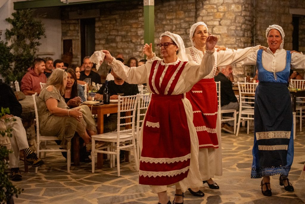 A lively evening event at ELaiolithos Naxos with local musicians playing the tsampouna and lyre, dancers in traditional Naxian folk costumes, and international travelers mingling and dancing together on a stone terrace