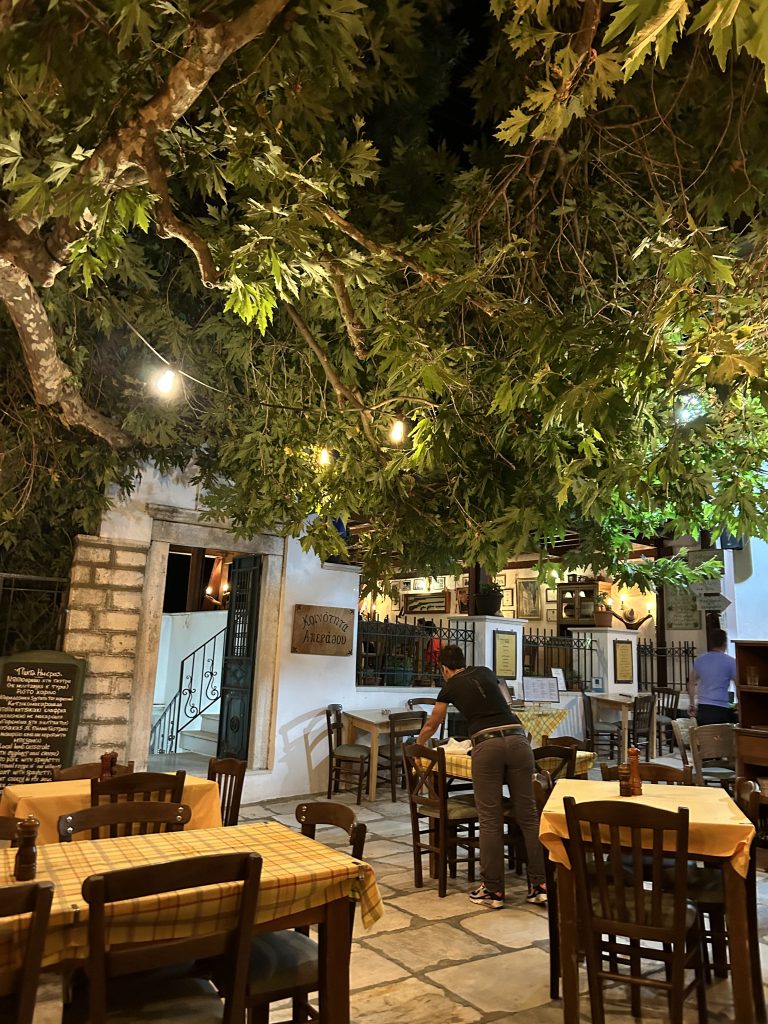 Authentic Greek village square on Naxos featuring traditional wooden taverna tables set under the shade of a massive, ancient plane tree with whitewashed stone paving and a peaceful mountain atmosphere.
