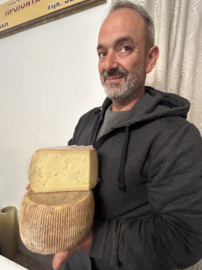 A 4th-generation Naxian shepherd and artisan cheesemaker holding a wheel of traditional Arseniko cheese in his shop in Chalki village
