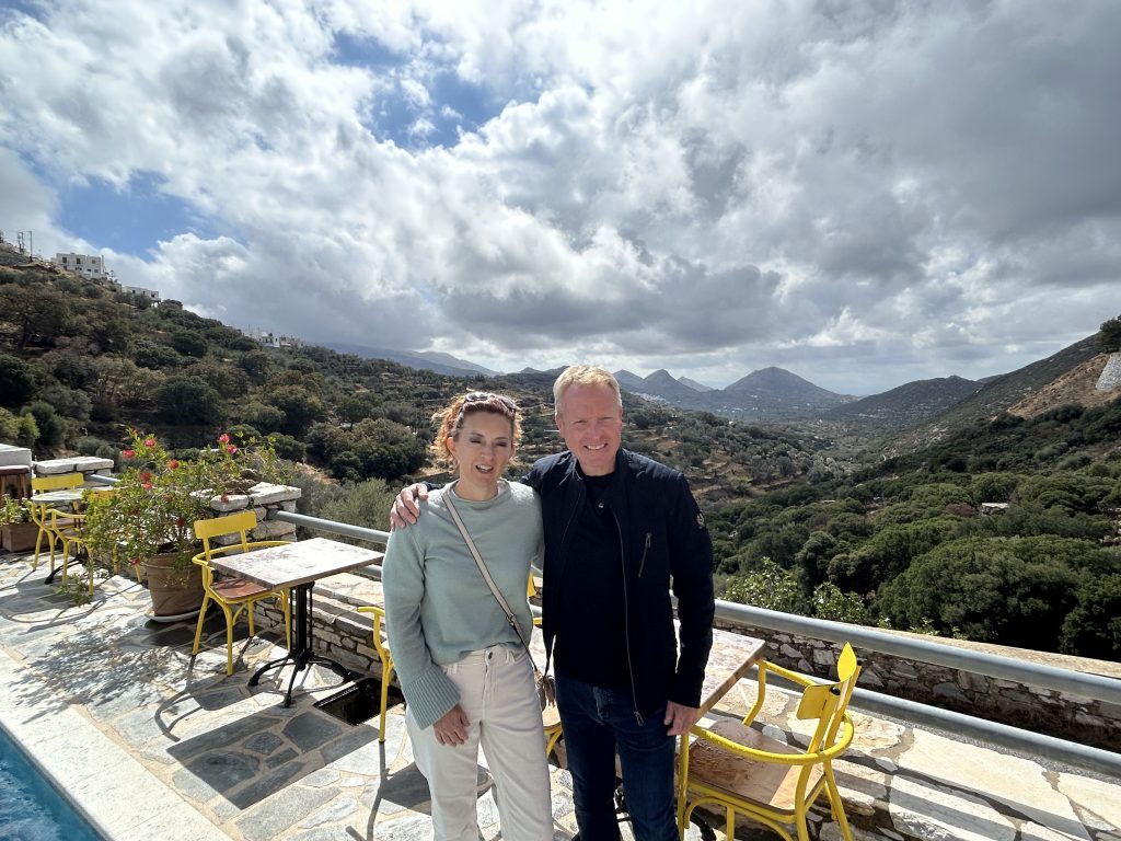A couple relaxing on a private terrace at ELaiolithos, enjoying expansive panoramic views of the green Naxian highlands and distant mountains.