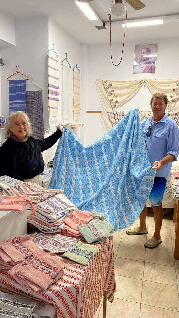 A master Naxian weaver with 65 years of experience working at a traditional wooden loom in her Chalki village exhibition, handcrafting intricate textiles for the ELaiolithos cultural ecosystem.